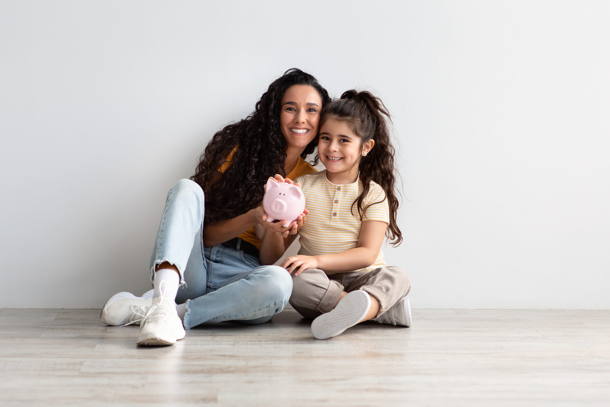 Financial Literacy. Portrait Of Young Mom And Her Little Daughter With Piggybank In Hands Sitting On Floor Near White Wall At Home, Smiling Mother Teaching Her Child Saving Money, Copy Space