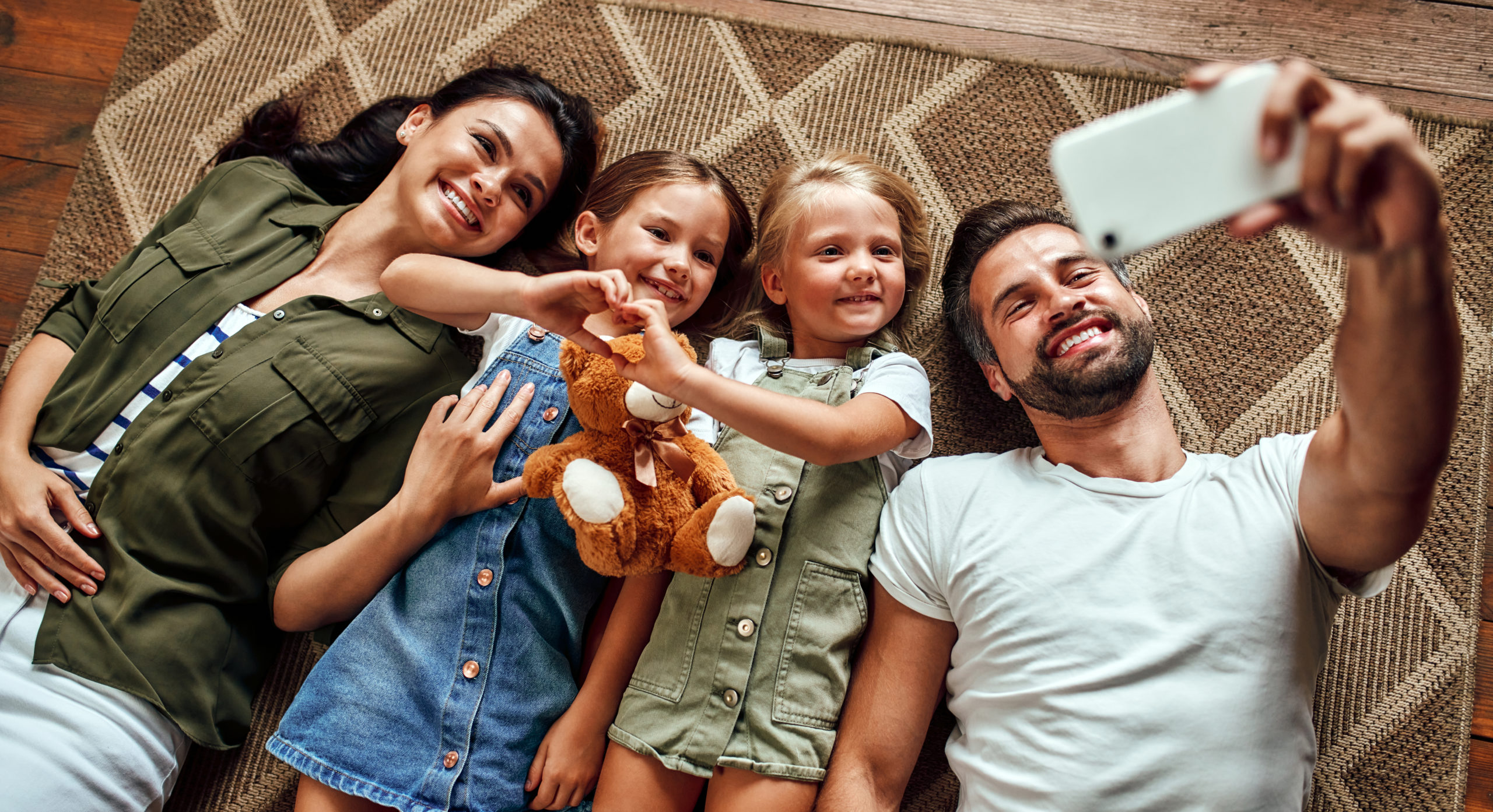 Dad, mom and two cute daughters lie on the carpet on the floor in the living room and take a selfie on a smartphone. Happy family have fun together.